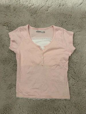 Ljusrosa topp med spetsdetalj - Supersöt ljusrosa topp från Monki med v-ringning, knappar framtill och vit spetskant vid halsen och nederkanten. Kortärmad.😊 skriv för fler frågor!