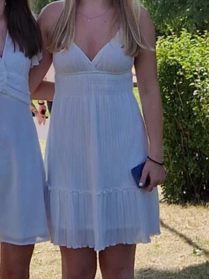 Vit kortklänning med smala axelband - Säljer en vit kortklänning med smala korsade axelband och v-ringad front. Klänningen har ett plisserat tyg och markerad midja som ger en snygg passform. Perfekt till sommaren och studenten eller skolavslutningen.