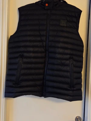 Svart dunväst från Moose Knuckles - Snygg svart dunväst från Moose Knuckles med quiltad design och två dragkedjeförsedda fickor framtill. Västen har hög krage och en diskret logotyp på bröstet. Perfekt för lager-på-lager och håller dig varm under kyliga dagar.