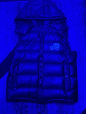 Svart dunväst från Moncler med huva - Snygg svart dunväst från Moncler med avtagbar huva och dragkedja framtill. Västen har två fickor med tryckknappar och Moncler-logga på bröstet. Perfekt för lager-på-lager och streetstyle.