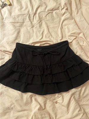Svart volangkjol med snörning - Säljer en svart kortkjol med volanger och snörning i midjan. Kjolen har flera lager volang som ger en cool och lekfull look. Perfekt för dig som gillar att sticka ut och vill ha något bekvämt och snyggt. Materialet känns mjukt och stretchigt.
