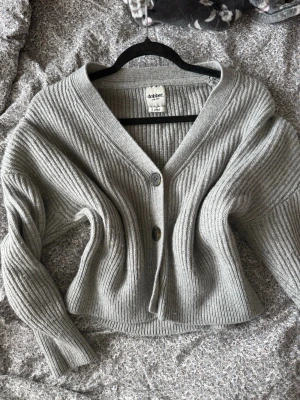 Grå ribbstickad kofta från Dobber - Grå ribbstickad kofta från Dobber med två knappar framtill och djup V-ringning. Koftan har långa ärmar och är tillverkad i ett mjukt, tjockare material som ger en avslappnad och trendig look.