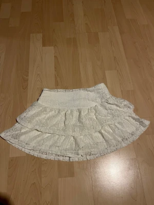 Vit volangkjol i spets - Söt vit kortkjol med volanger och spetsdetaljer. Kjolen har en feminin och romantisk look med två lager volanger och bred midja. Perfekt för dig som gillar en ljus och somrig stil.