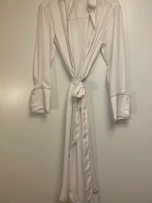 Vit omlottklänning med knytband - Vit omlottklänning med långa ärmar, markerade manschetter och klassisk krage. Klänningen har knytband i midjan och är tillverkad i ett mjukt tyg. Perfekt för dig som gillar en stilren och enkel look.