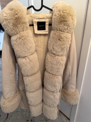 Beige pälsjacka från Capone - Säljer en fluffig beige pälsjacka från Capone med stora, mjuka fuskpälsdetaljer längs krage, ärmslut och framkant. Jackan har en rak passform och är helfodrad med samma mjuka material på insidan. Perfekt för kalla vinterdagar.