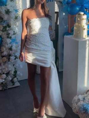 Vit axelbandslös klänning - Snygg vit axelbandslös klänning med kort framsida och lång släp baktill. Överdelen har spetsdetaljer och kjolen är i satinliknande material. Perfekt för studentfirandet och ger en elegant look.