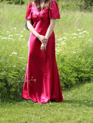 Röd långklänning - Snygg röd långklänning i satin med v-ringning och puffiga kortärmar. Klänningen har en elegant rosettdetalj framtill och ett glansigt material som ger en lyxig känsla. Perfekt för dig som vill sticka ut och känna dig extra glamorös. Jag är 157cm för referens