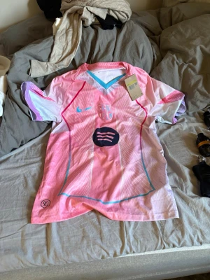 Barca tröja helt ny - Säljer en rosa och vit fotbollströja med blå och röda detaljer. Tröjan har korta ärmar, V-ringning och tryck på bröstet samt logga från märket 90. Materialet är lätt och andas, perfekt för träning eller match.