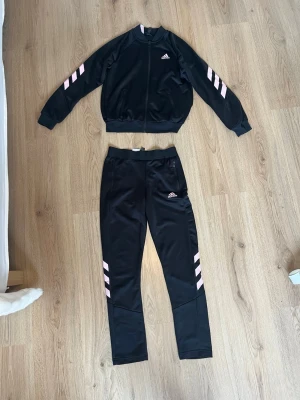 Svart Adidas träningsset med rosa detaljer - Säljer ett svart set  från Adidas med matchande jacka och byxor. Setet har rosa ränder på ärmar och ben. Nästan aldrig andvända. Ny pris jacka runt 300-500 byxor 250-450. Priset kan diskuteras.💞