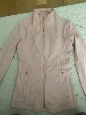 lululemon define jacket - Snygg ljusrosa jacka från Lululemon med figurnära passform och hel dragkedja framtill. Jackan har två fickor med dragkedja och hög krage. Perfekt för lager-på-lager och en sportig look.