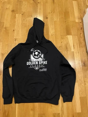 Gildan Svart hoodie Golden Spike Classic stl M  - Svart hoodie med tryck 'Golden Spike Classic' och en fotbollsgrafik på bröstet. Huva med dragsko och stor magficka framtill. Perfekt för dig som gillar sportiga och bekväma plagg. Materialet känns mjukt och skönt mot huden.