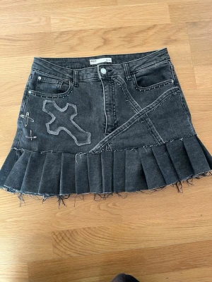 Svart minidenimkjol med unika detaljer  - Cool svart denimkjol  med råa kanter, plisserad nederdel, korsapplikation och säkerhetsnålar som detaljer. Kjolen har en unik design med synliga sömmar och fickor fram. Perfekt för dig som gillar edgy streetstyle.