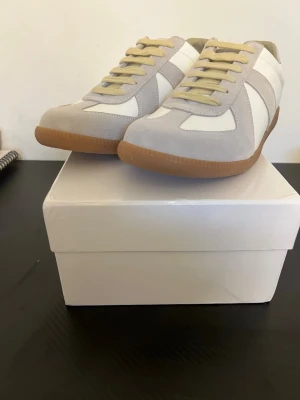 Maison Margiela skor  - Snygga sneakers i vitt och ljusgrått med beige snörning och klassisk gummisula. Ovandelen är i en mix av mocka och slätt material, perfekt för en clean och stilren look. Passar dig som gillar minimalistisk design och neutrala färger. Av märket Maison Margiela somriga med rik stil