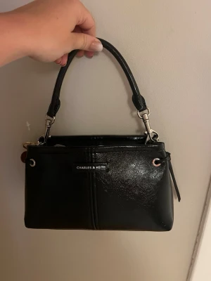 Svart handväska från Charles & Keith - Snygg svart handväska från Charles & Keith i glansigt konstläder. Väskan har ett kort handtag, silverfärgade metalldetaljer och dragkedja upptill. Insidan har ett fack med dragkedja och logotypen syns både utvändigt och invändigt.