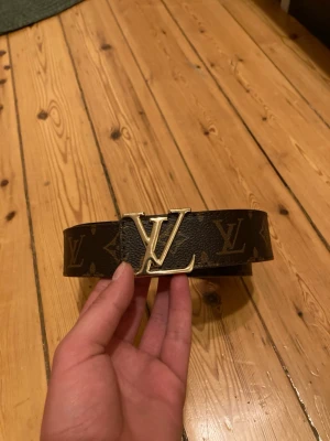 Märkes Bälte monogram skärp - Snyggt skärp i mörkbrunt skinn med klassiskt monogrammönster och stor logga i guld framtill. Bältet har en exklusiv känsla och passar perfekt till att lyfta vilken outfit som helst.