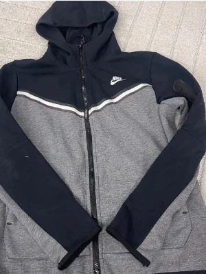 Nike hoodie med dragkedja svart/grå - Nike hoodie i svart och grått med hel dragkedja framtill och vit logga på bröstet. Hoodien har en huva, två fickor och en vit rand över bröstet. Tillverkad i mjukt sweatshirtmaterial, perfekt för en sportig och avslappnad stil.