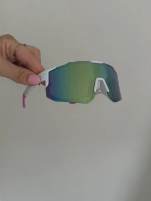 Sportiga vita solglasögon med spegelglas sportglasögon  - Säljer ett par sportiga solglasögon med vit båge och stora spegelglas i grönt och blått. Glasögonen har en modern, kantig form och passar perfekt för en aktiv livsstil. Snygg detalj med rosa på skalmarna. Perfekta för dig som gillar att sticka ut.