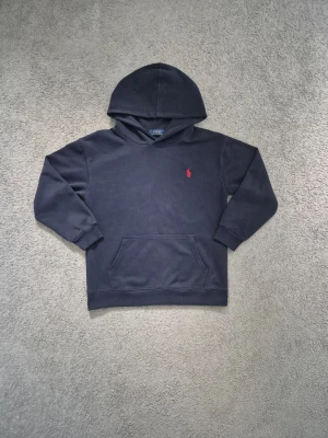 Polo Ralph Lauren - Mörkblå hoodie från Polo Ralph Lauren med klassisk känguruficka och huva. Broderad röd logga på bröstet ger en stilren touch. Mjuk bomullsblandning och ribbade muddar vid ärmslut och nederkant. Perfekt för en avslappnad och snygg look.