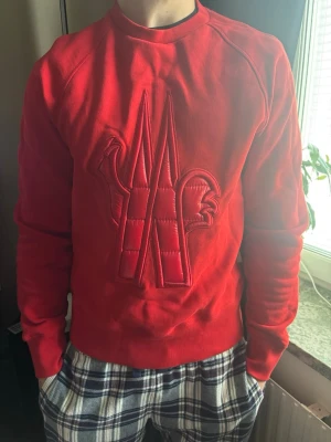 Röd Moncler sweatshirt med broderi - Cool röd sweatshirt från Moncler med stort broderat och vadderat logomärke på bröstet. Tröjan har rund halsringning och långa ärmar. Perfekt för dig som vill sticka ut med en exklusiv streetwear-look.