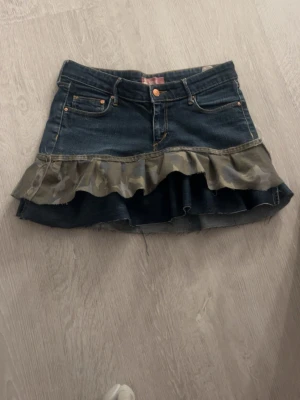 Jeanskjol med volang och camouflage - Cool jeanskjol i mörkblå denim med volangkant i camouflage-mönster. Kjolen har klassiska fickor fram och bak samt råa kanter nertill för en trendig look. Perfekt för dig som vill sticka ut med en unik stil.  Jag har sytt den själv från skinny jeans. Midjemått är ca 39 cm och längden ca 32cm. 