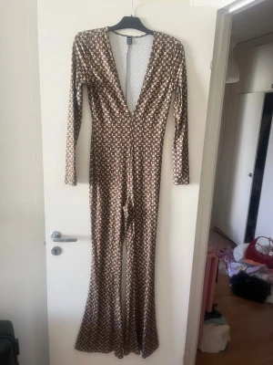 Mönstrad jumpsuit med djup V-ringning - Säljer en långärmad jumpsuit från Boohoo med brunt och beige retroinspirerat mönster. Djup V-ringning framtill och utsvängda ben ger en snygg siluett. Stretchigt material som sitter tajt och bekvämt. Perfekt för dig som gillar att sticka ut med din stil.