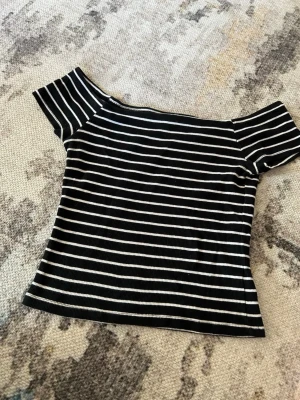 Randig offshoulder topp svart/vit - Snygg ribbad offshoulder topp med svarta och vita horisontella ränder. Toppens passform är tight och den är kortärmad, perfekt för en trendig look. Materialet känns mjukt och stretchigt. Det står L men passar som S / XS