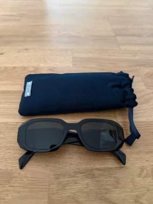 Svarta solglasögon från Prada - Snygga svarta solglasögon från Prada med rektangulära bågar och mörka glas. Klassisk och stilren design med breda skalmar där Prada-loggan syns tydligt på sidan. Kommer med ett fodral. Kvitto finns.