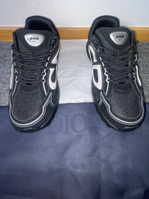 Svarta Dior B30 sneakers - Svarta Dior B30 sneakers med silvriga detaljer och reflekterande inslag. Skorna har en sportig design med mesh och syntetmaterial, samt tydlig Dior-logga på sidan. Klassisk snörning och robust sula för en modern look.