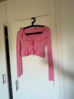 Rosa croppad topp med volang - Söt rosa croppad topp med långa ärmar och volangdetaljer längs ringningen och framtill. Toppen har knyt framtill och en v-ringad urringning som ger en feminin look. Perfekt för dig som gillar trendiga och romantiska plagg.