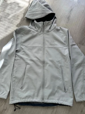 Ljusgrå vindjacka från Arc'teryx - Snygg ljusgrå vindjacka från Arc'teryx med huva och dragkedja framtill. Jackan har två praktiska fickor med dragkedja och broderad logga på bröstet. Tillverkad i ett slätt, vindtätt material som passar perfekt för blåsiga dagar.