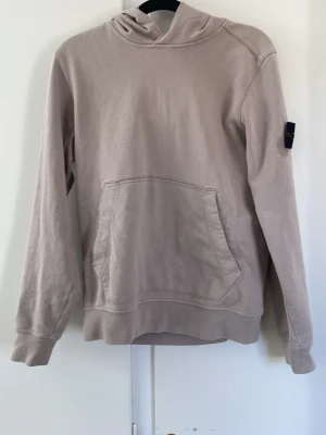 Beige hoodie från Stone Island - Säljer en beige hoodie från Stone Island med klassisk patch på ärmen. Hoodien har en stor magficka och en avslappnad passform. Perfekt för dig som gillar streetwear och vill ha något stilrent men ändå unikt. Materialet känns mjukt och skönt mot huden.