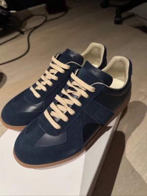 Blå Maison Margiela Gats - Snygga blå Maison Margiela Replica sneakers med ovandel i mocka och läder, beige snörning och vit insida. Skorna har en klassisk låg siluett och gummisula som ger en retrovibe. Perfekta för dig som gillar stilrena och tidlösa sneakers.