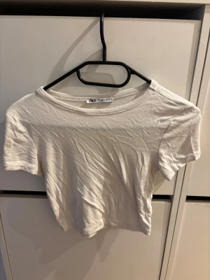 Vit croppad t-shirt från Zara - Säljer en enkel och stilren vit croppad t-shirt från Zara. T-shirten har korta ärmar, rund halsringning och är gjord i mjuk bomull. Perfekt basplagg att matcha med allt i garderoben.
