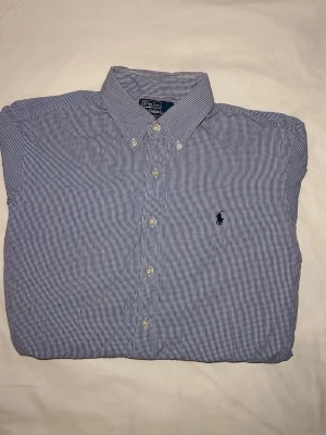 Ralph Lauren | Skjorta | L - Klassisk blårutig skjorta från Polo Ralph Lauren med button-down krage och broderad logga på bröstet. Skjortan har lång ärm, vita knappar och är tillverkad i bomull. Perfekt för en stilren look. Mått: 71 Längd, 52 Bredd