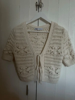 Virkat kofta från Zara i offwhite - Superfin virkad kofta från Zara i offwhite med kort ärm och knytning framtill. Koftan har ett luftigt mönster med små blommor och bred ribbad kant nedtill och vid ärmslut. Perfekt att slänga över ett linne eller klänning.