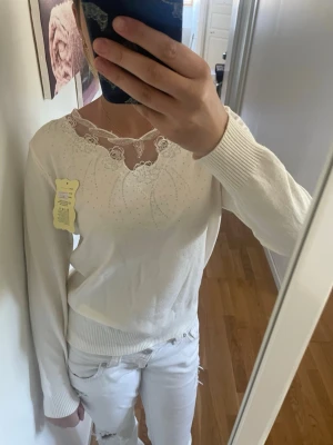 Vit tröja med spetsdetalj - Snygg vit tröja med lång ärm och vacker spets runt halsringningen. Tröjan har diskreta glittriga detaljer framtill och ribbade muddar vid ärmslut och nederkant. Perfekt för dig som gillar en stilren look med en twist.