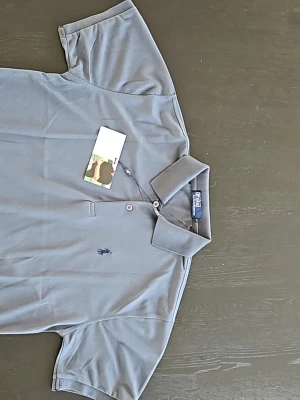 Grå pikétröja från Polo Ralph Lauren - Snygg grå pikétröja från Polo Ralph Lauren med klassisk krage och två knappar framtill. Tröjan har korta ärmar och den ikoniska broderade loggan på bröstet. Perfekt för en stilren och avslappnad look.