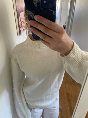 Vit stickad tröja med pärlor - Säljer en vit stickad tröja med pärldetaljer på axlar och ärmar. Tröjan har en hög ribbad krage och långa ärmar med ribbade muddar. Perfekt för dig som gillar en stilren look med lite extra detaljer.