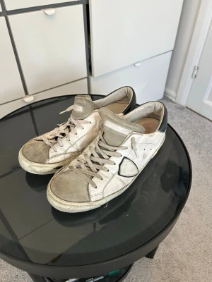 Vita sneakers från Philippe Model - Säljer ett par vita Philippe Model sneakers med grå detaljer och mörkblå häl. Skorna har klassisk låg profil, vita snören och loggan på sidan. Ovandelen är i läder och mocka, och sulan är i gummi. Skorna har stått utan användning nu, men användes en hel del förra sommaren. Man ser på dem att de är användna men skicket är fortfarande bra. Hör gärna av er