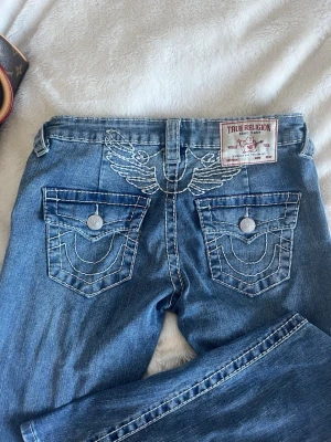 True Religion blå jeans med brodyr - Säljer ett par blå jeans från True Religion med ikonisk vit brodyr på bakfickorna och ovanför, samt tydliga kontrastsömmar. Klassisk femficksmodell med knappstängning och patch med logga bak i midjan. Perfekta för en trendig streetstil.