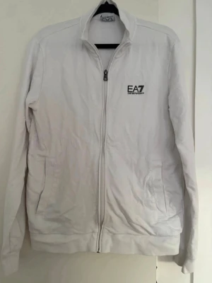 Vit zip-hoodie från EA7 Emporio Armani - Snygg vit hoodie med dragkedja från EA7 Emporio Armani. Tröjan har hög krage, två fickor framtill och svart logga på bröstet. Tillverkad i mjukt material som känns skönt mot huden. Perfekt för en clean och stilren look.
