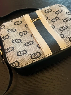 Beige axelväska från Liu Jo - Snygg beige axelväska från Liu Jo med svart och vit rand på framsidan samt guldfärgade detaljer. Väskan har ett monogrammönster i svart och justerbar axelrem i svart konstläder. Perfekt för dig som gillar trendiga accessoarer med logga.