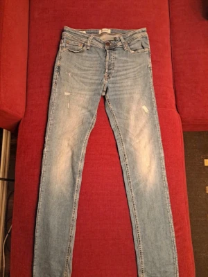 Ljusblå jeans Jack & Jones - Säljer ett par ljusblå jeans från Jack & Jones med slitna detaljer och klassisk femficksmodell. Jeansen har dragkedjegylf och är tillverkade i bomull med lite stretch för skön passform. Perfekta till en avslappnad look.