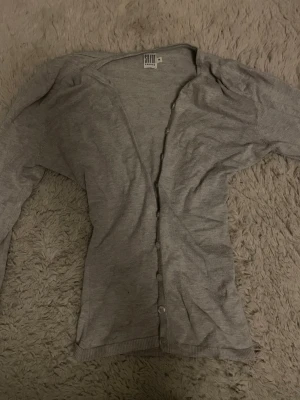 Ljusgrå tunn kofta med knappar - Ljusgrå tunn kofta med långa ärmar och knäppning framtill. Sitter tajt vid midjan då jag har sytt in den. Enkel och stilren design, perfekt att slänga över en t-shirt. Ribbstickade muddar vid ärmslut och nederkant.