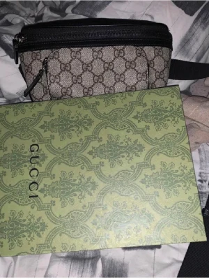 Gucci axelväska med GG-mönster - Snygg axelväska från Gucci i beige canvas med klassiskt GG-monogram och svarta detaljer. Väskan har justerbart svart band och dragkedja upptill. Perfekt för att bära det viktigaste med stil.