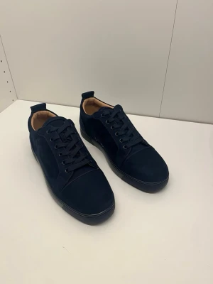 Mörkblå sneakers från Christian Louboutin - Snygga mörkblå sneakers i mocka från Christian Louboutin med klassisk låg siluett och ikoniska röda sulor. Skorna har svarta skosnören och diskreta detaljer, samt en mjuk innersida i beige läder. Perfekta för dig som vill sticka ut med stil.