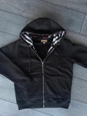Burberry zip upp - Svart hoodie från Burberry med dragkedja framtill och klassiskt rutigt foder i huvan. Tröjan har två fickor, ribbade muddar och snören i huvan. Perfekt för en stilren och avslappnad look.
