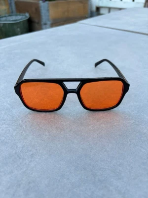 Svarta solglasögon med orange glas - Coola solglasögon med svart ram och rektangulära orange glas. Snygg detalj med unik brygga mellan glasen. Perfekta för dig som vill sticka ut med en retrovibe. Lätta och bekväma att bära hela dagen. Perfekt nu till sommaren ☀️