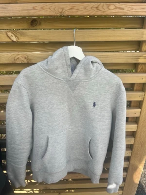 Grå hoodie från Polo Ralph Lauren - Snygg grå hoodie från Polo Ralph Lauren med klassisk huva och känguruficka framtill. Diskret broderad logga i mörkblått på bröstet. Är egentligen storlek M men passar bäst för S-XS! 