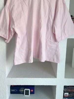 Ljusrosa blus med en söt liten rosett i nacken - Säljer en ljusrosa blus i bomull med v-ringning och korta ärmar. Toppen har en avslappnad passform och är perfekt för en chill och stilren look. Enkel design utan mönster eller detaljer, vilket gör den lätt att matcha med olika outfits.
